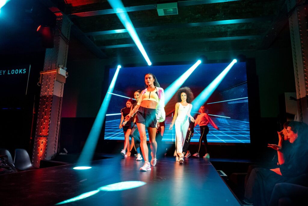fashion show, runway, models, sportswear, athletic wear, catwalk, stage lights, futuristic, urban fashion, audience, performance, LED screen, event, fashion week, indoor venue